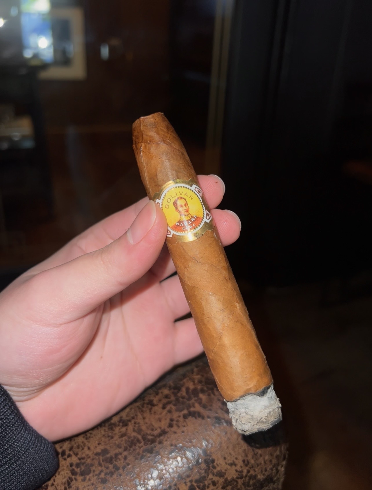 A Bolivar Belicosos Finos cigar held in hand, with its gold band and ash visible
