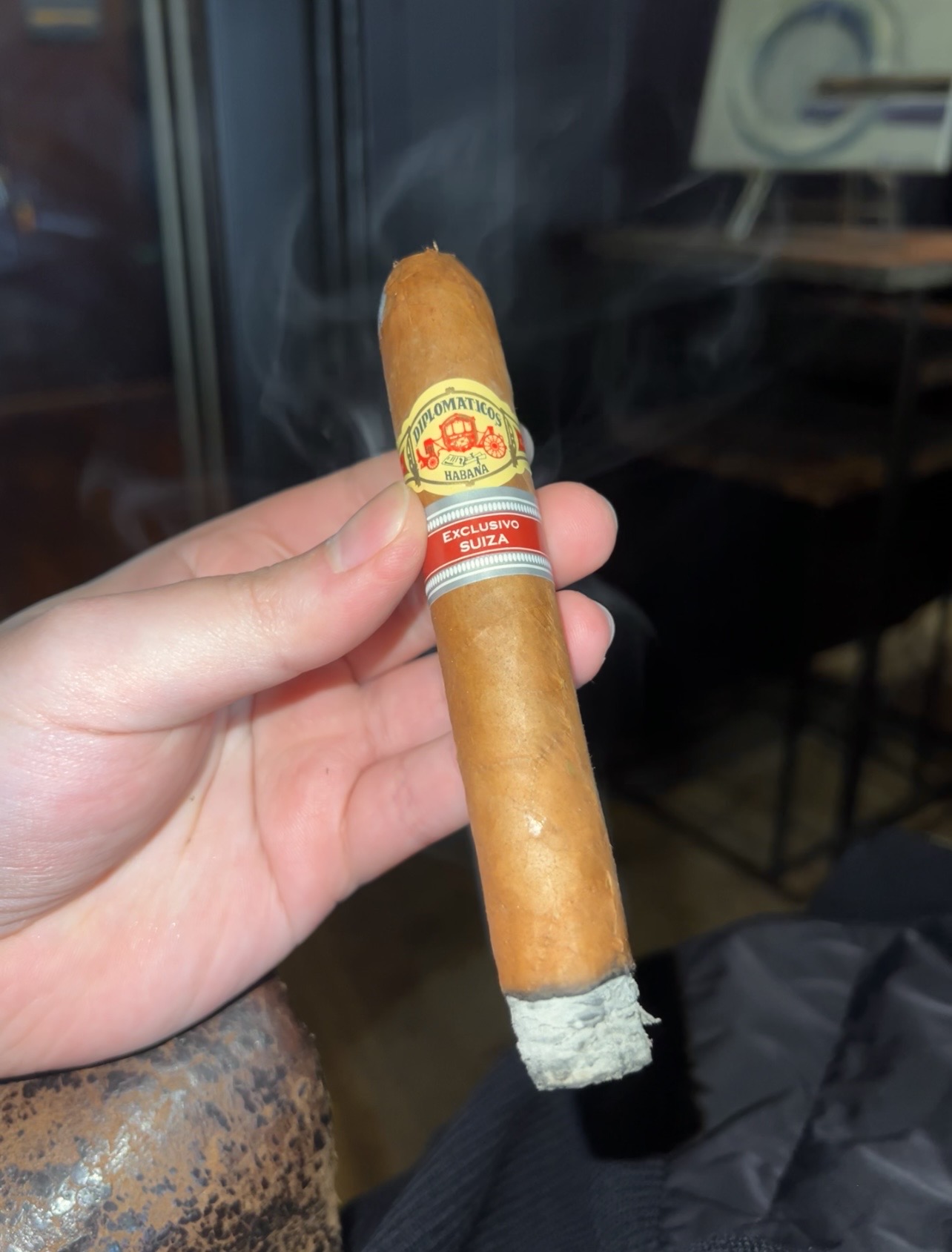 A Diplomáticos El Emisario cigar held in hand, showing its yellow band, red Exclusivo Suiza band, and ash