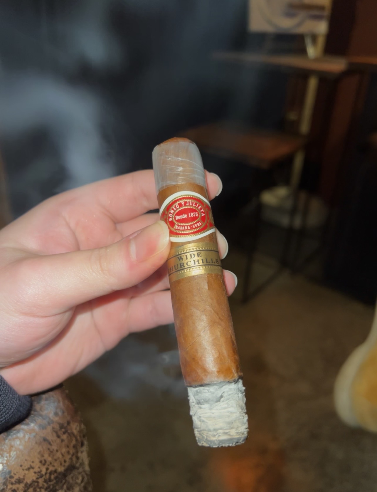 A Romeo y Julieta Wide Churchills cigar held in hand, showing its red and gold bands and ash