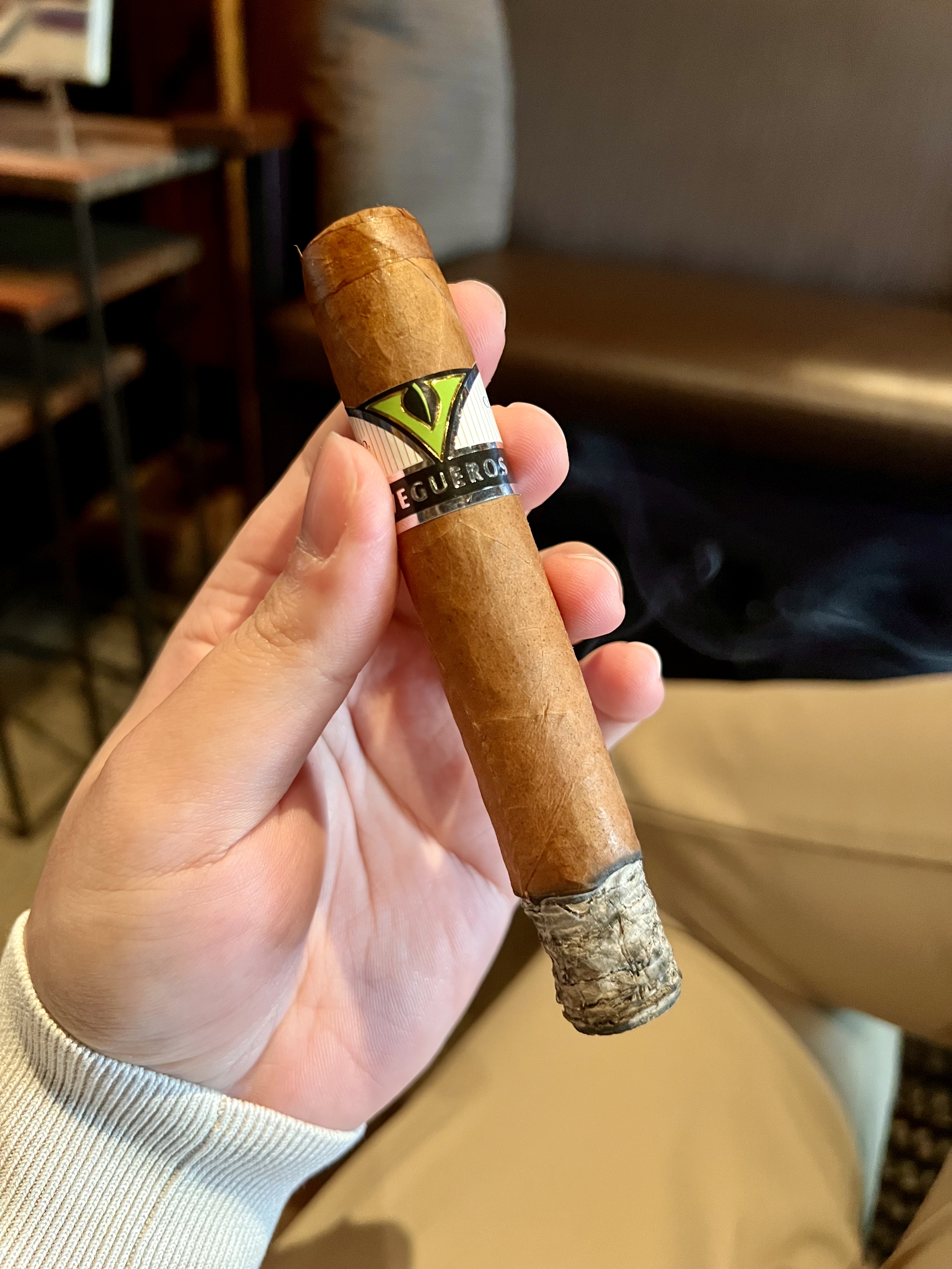 A hand holding a lit Vegueros Tapados cigar with a green and silver band and a long ash